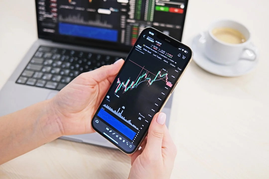 Mão de uma pessoa segurando um smartphone exibindo um aplicativo financeiro com gráficos de ações e portfólio de investimentos, com um laptop e documentos financeiros ao fundo.